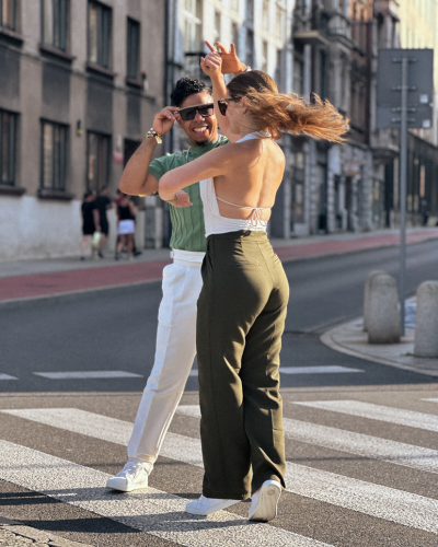 Ein Paar tanzt fröhlich auf einem Zebrastreifen in der Stadt, im Hintergrund sind Gebäude zu sehen.