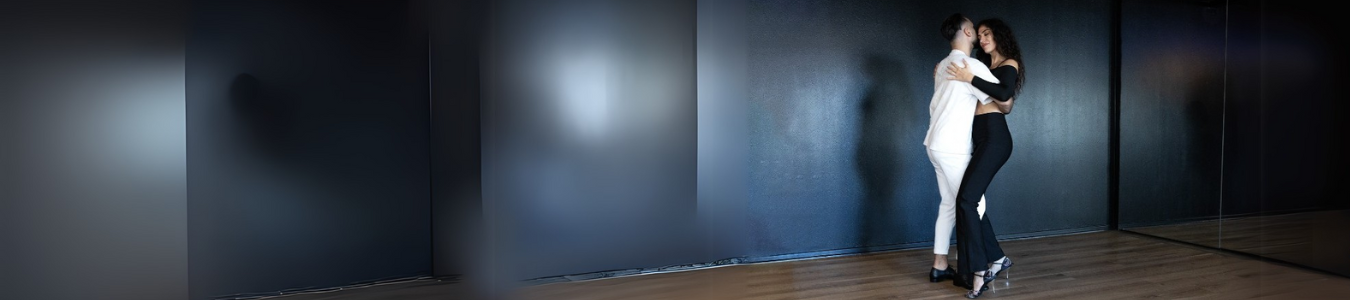 Zwei Menschen tanzen eng beieinander in einem schwach beleuchteten Studio, ihre Spiegelbilder sind im Spiegel sichtbar.