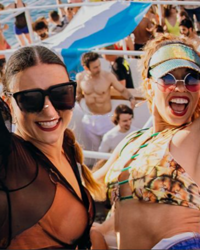 Zwei Frauen lächeln bei einer lebhaften Party im Freien, mit Menschen und einer großen Flagge im Hintergrund.