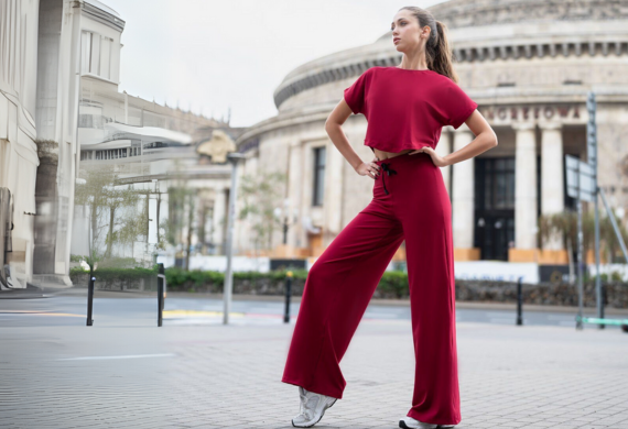 Frau im roten Outfit posiert selbstbewusst auf einer Stadtstraße mit verschwommenem historischen Gebäude im Hintergrund.
