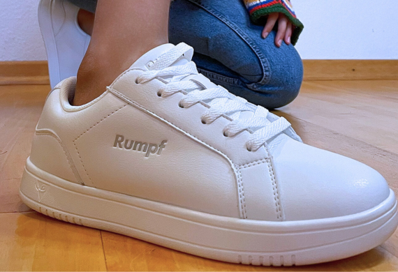 Nahaufnahme von weißen Turnschuhen mit dem Markennamen „Rumpf“ auf Holzboden, im Hintergrund kniende Person in Jeans.