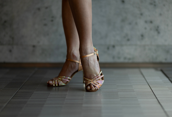 Nahaufnahme einer Person in glitzernden Riemchentanzschuhen auf einem gefliesten Boden vor einer strukturierten grauen Wand im Hintergrund.