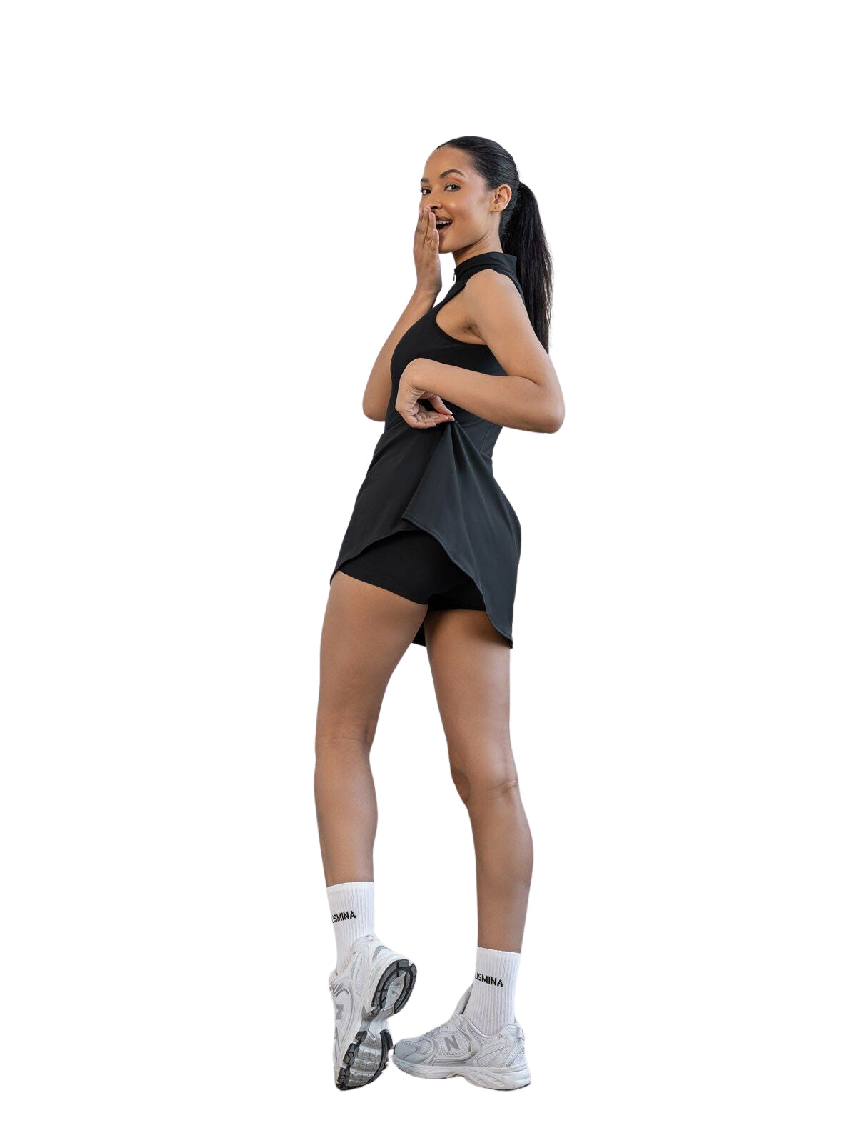 Frau trägt Lismina 3050 Jumpsuit in Schwarz, dazu weiße Socken und Sneaker, lächelt mit Hand am Gesicht.