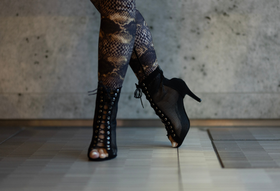 Person in Leggings mit Schlangenhautmuster und hochhackigen Schnürstiefeln steht auf einem Fliesenboden.