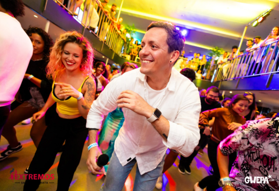 Menschen tanzen energisch in einer lebhaften, farbenfrohen Partykulisse, während ein Mann die Bühne führt und ein Mikrofon hält.