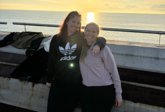 Zwei Frauen stehen Arm in Arm an einem Geländer bei Sonnenuntergang und lächeln, im Hintergrund ist das Meer zu sehen.