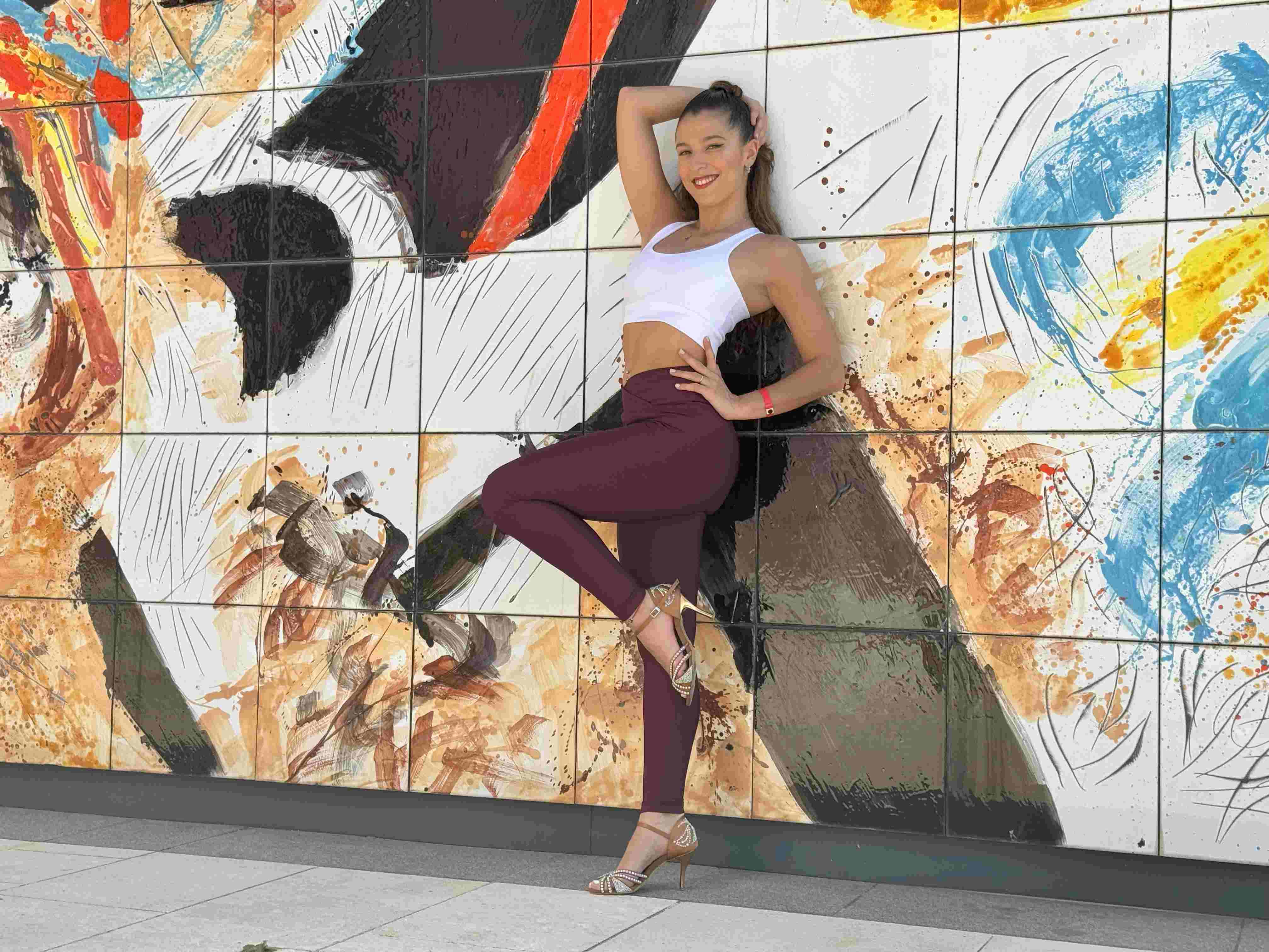 Person, die vor einem farbenfrohen abstrakten Wandgemälde posiert und ein weißes Oberteil, kastanienbraune Leggings und High Heels trägt.