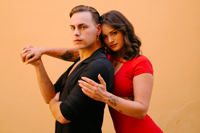 Zwei Personen posieren eng beieinander vor einer pfirsichfarbenen Wand, eine in schwarzem und eine in rotem Outfit.