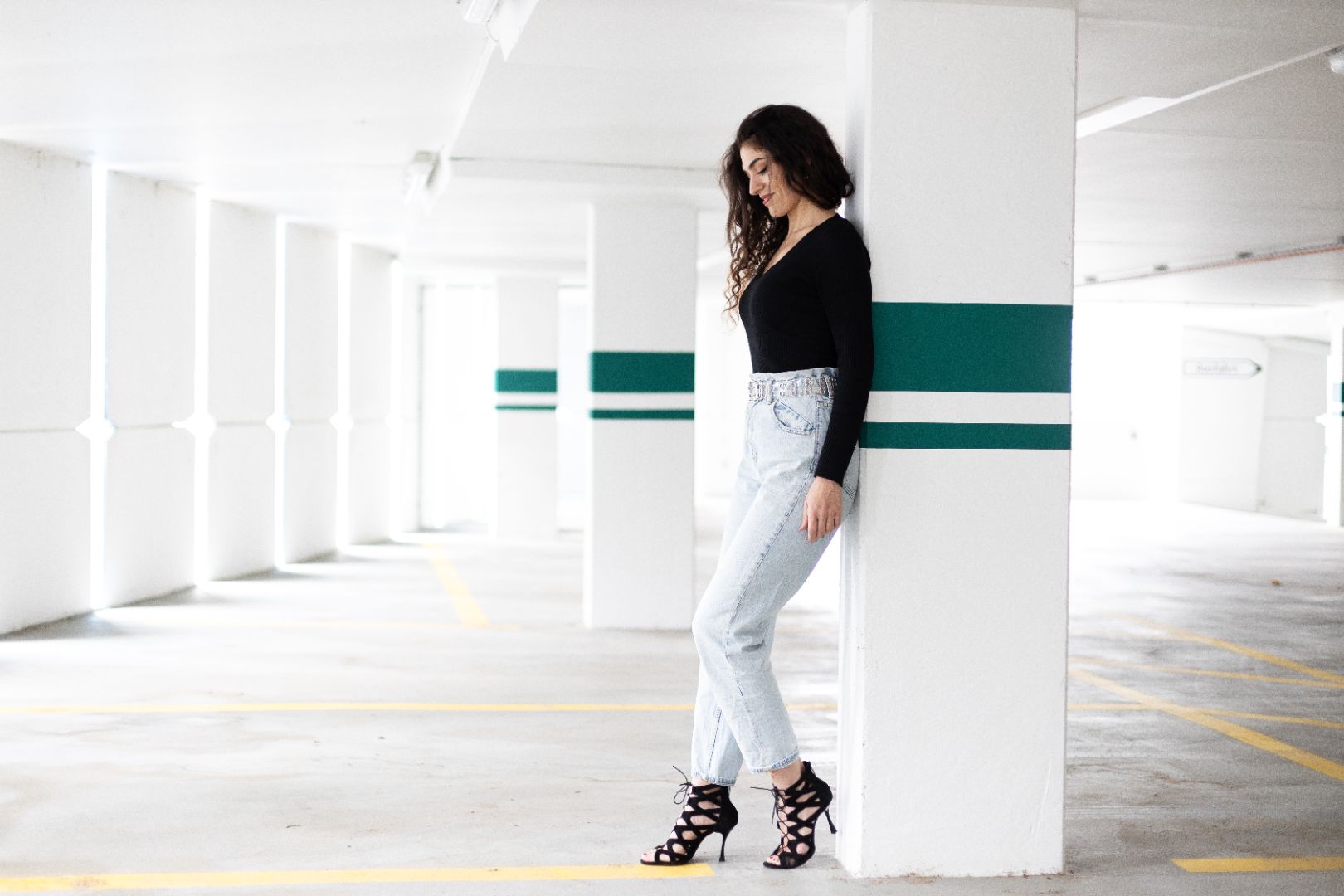 Frau in schwarzem Top und Jeans lehnt an einer Säule in einem hellen Parkhaus.