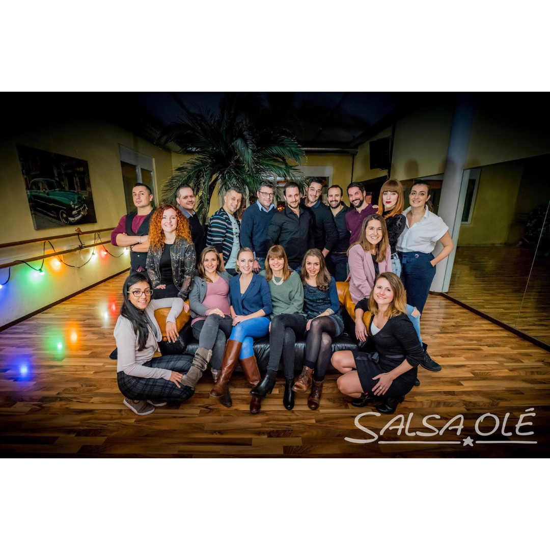 Eine Gruppe posiert in einem Tanzkurse-Studio auf einem Boden mit der Aufschrift „SalsaOlé“.