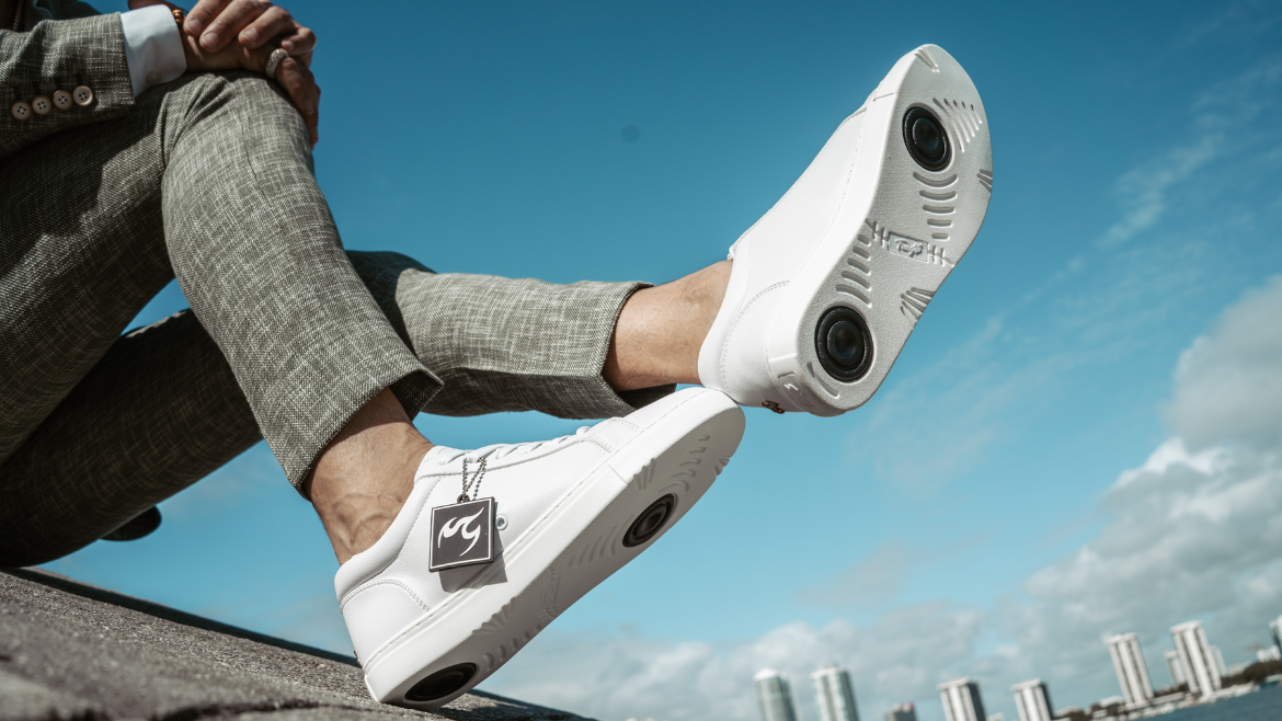 Eine Person in grauer Hose trägt weiße Turnschuhe mit einem einzigartigen Sohlendesign und sitzt vor dem Hintergrund eines blauen Himmels.