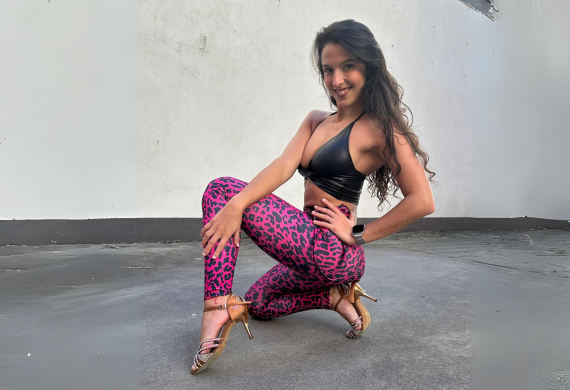 Frau in schwarzem Top und rosa gemusterten Leggings posiert mit einem angewinkelten Knie und trägt High Heels vor einem schlichten Hintergrund.