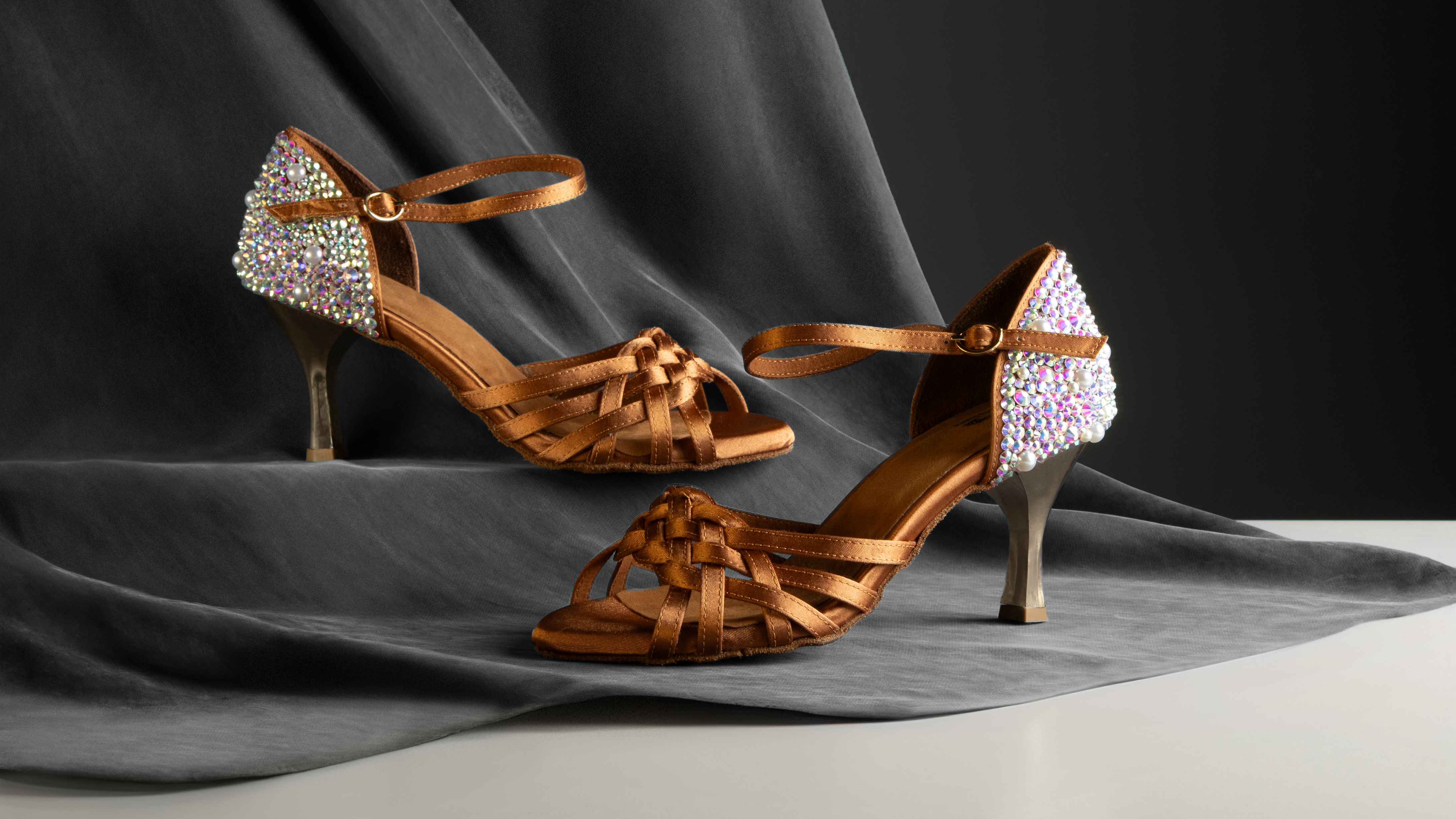 Braune Riemchensandalen mit strassbesetztem Absatz auf grauer Stoffoberfläche.