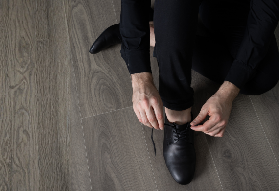 Auf einem Holzboden kniende Person, die die Schnürsenkel eines schwarzen Schuhs bindet und schwarze Kleidung trägt.
