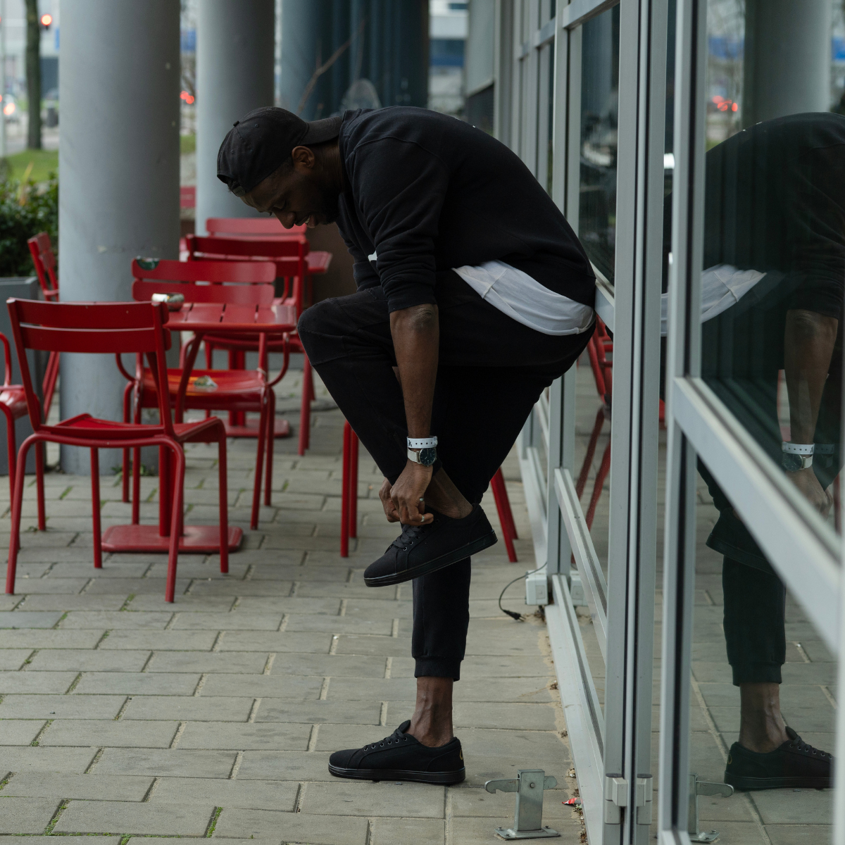 Mann, der an einem Fenster lehnt und dabei Schnürsenkel bindet, im Hintergrund rote Stühle und Tische.