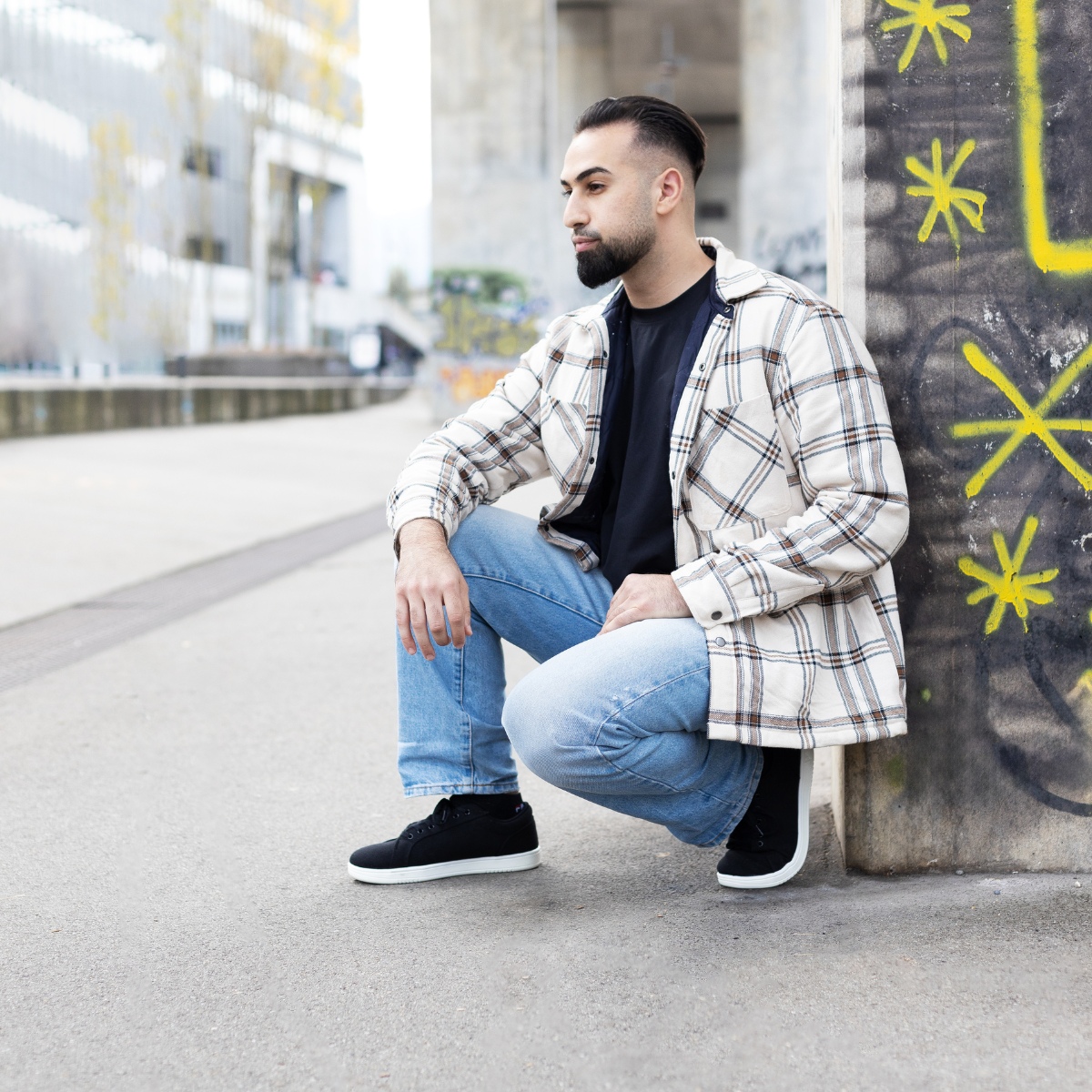 Mann in kariertem Hemd und Jeans kauert an einer Graffiti-Wand in städtischer Umgebung.