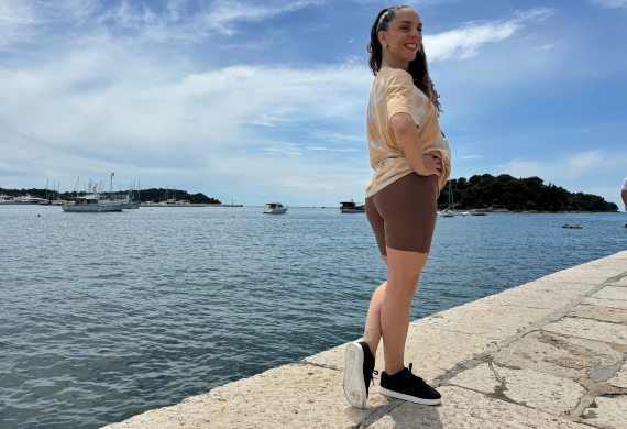 Lächelnde Person am Meer, mit Booten auf dem Wasser und einem teilweise bewölkten Himmel darüber.