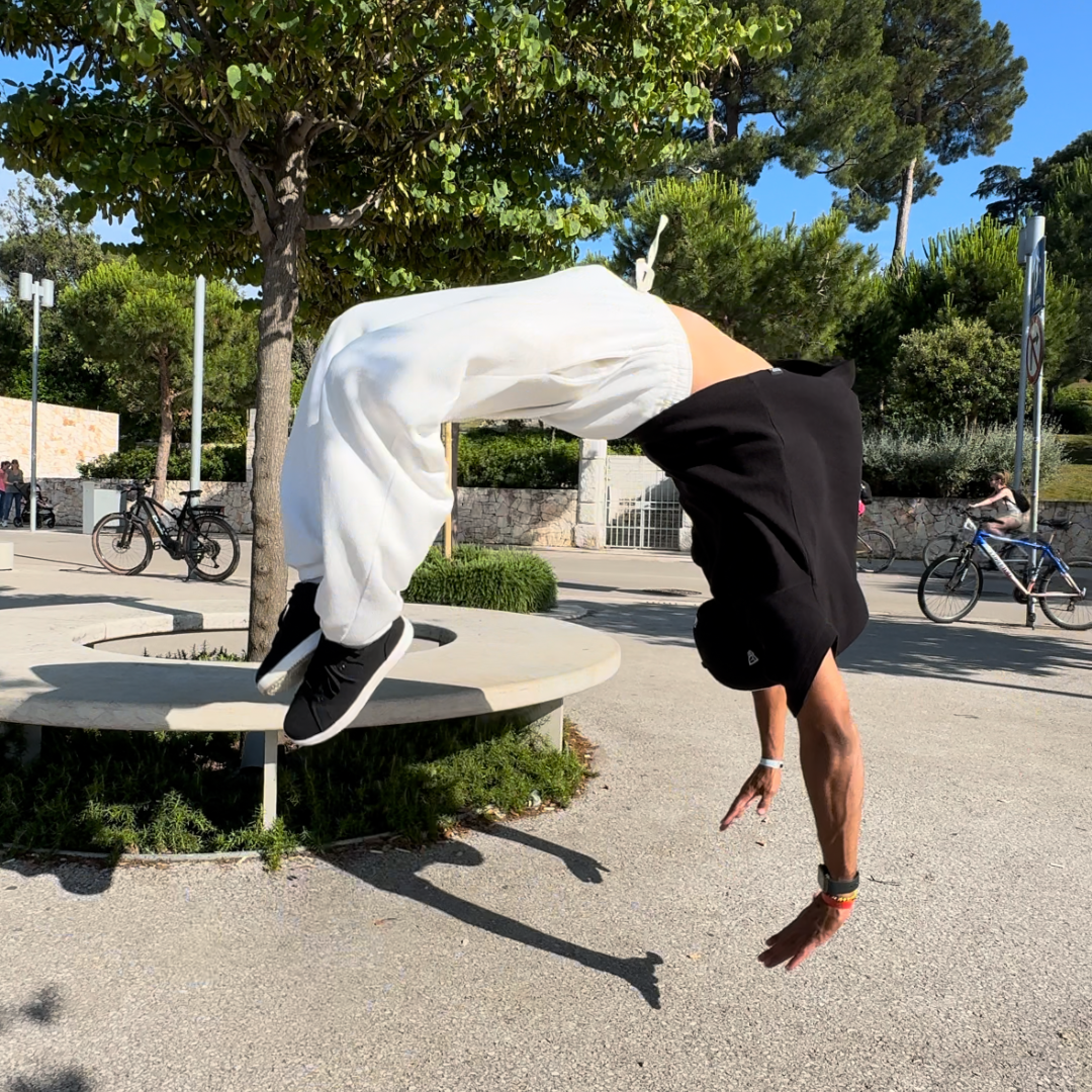 Person in der Luft, die in der Nähe eines Baums in einem Park einen Rückwärtssalto macht; sie trägt ein schwarzes Hemd, weiße Hosen und schwarze Schuhe.