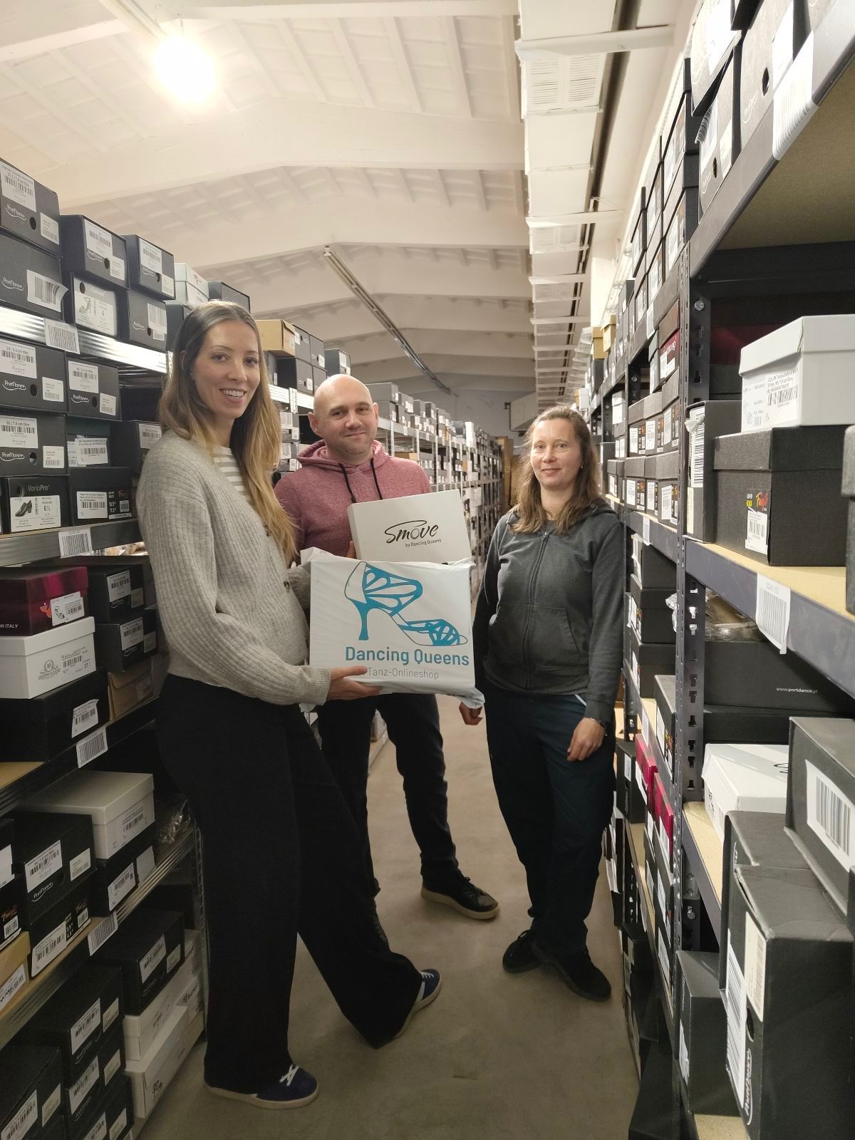 Drei Personen stehen in einem Schuhlager und halten eine Kiste mit der Aufschrift „Dancing Queens“.