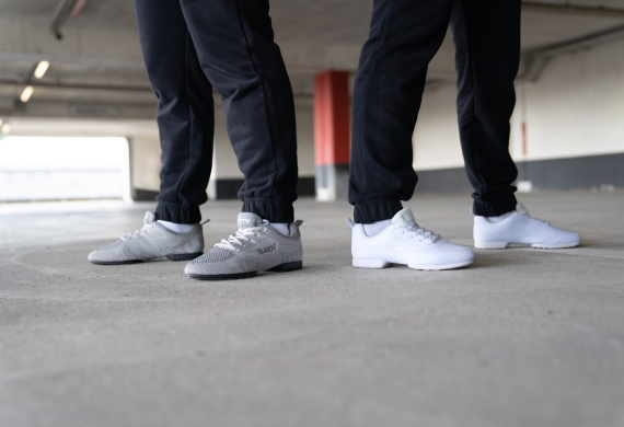 Zwei Personen in schwarzen Hosen und weißen Sportschuhen stehen auf einem Betonboden in einem Parkhaus.