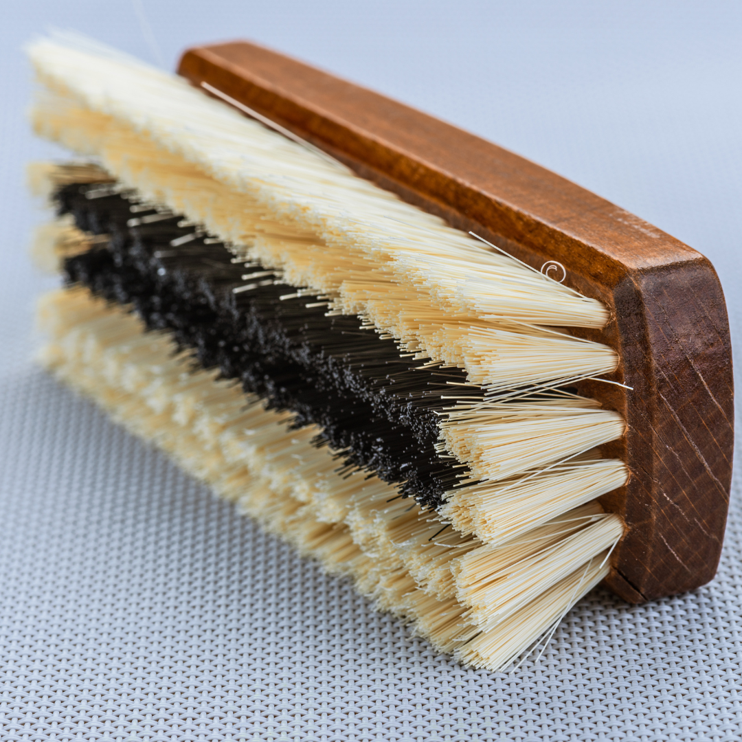 Nahaufnahme einer Scheuerbürste mit Holzgriff und zweifarbigen Borsten auf einer grau strukturierten Oberfläche.