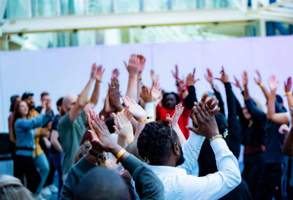 Eine bunt gemischte Gruppe von Menschen, die bei einer Indoor-Veranstaltung enthusiastisch die Hände heben und klatschen.