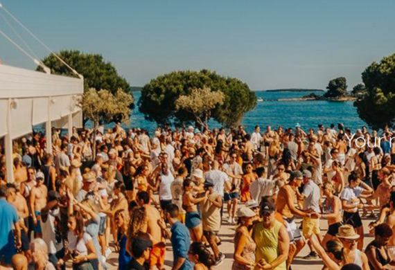 Eine große Menschenmenge versammelt sich und tanzt an einem sonnigen Tag im Freien am Meer, mit Bäumen und blauem Wasser im Hintergrund.