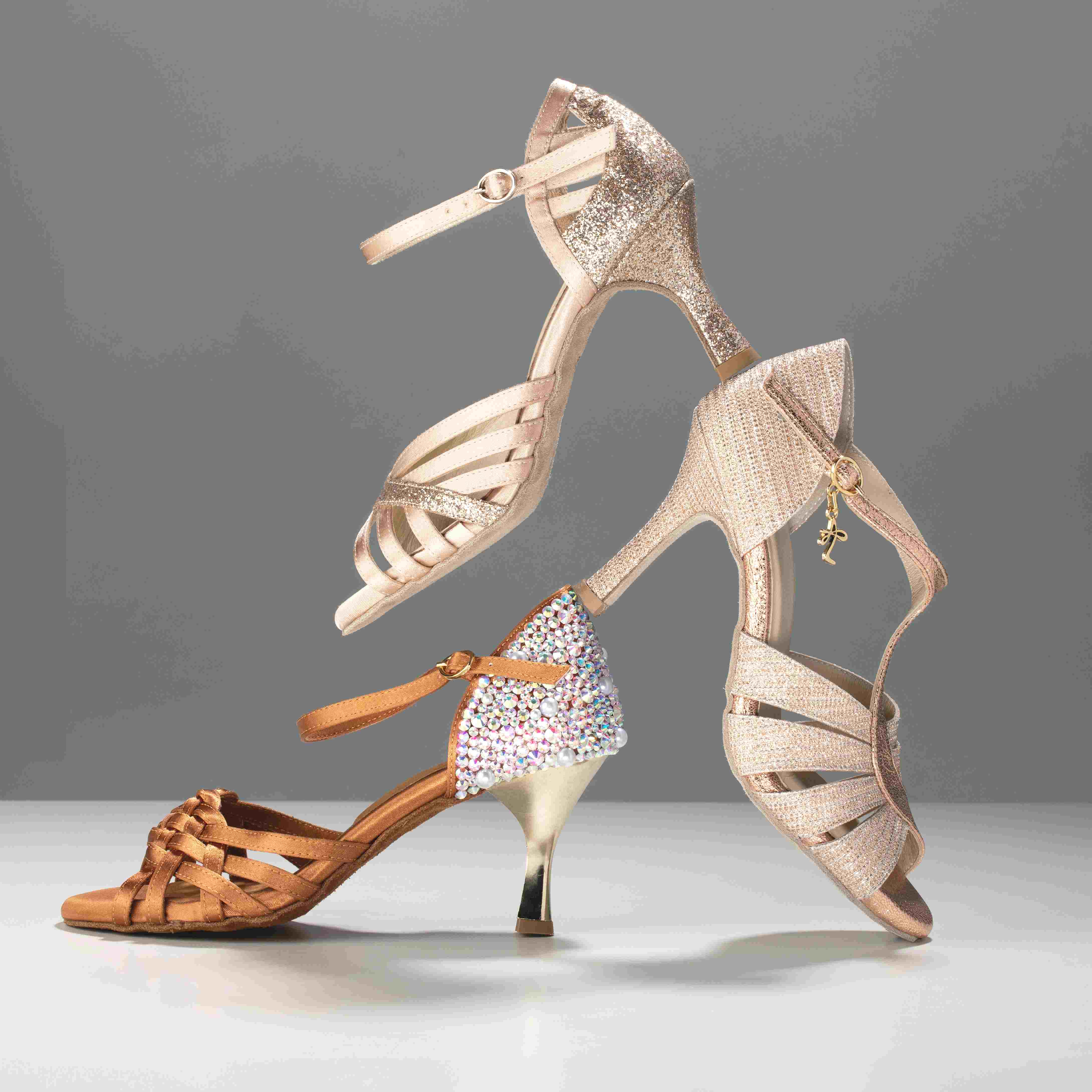 Drei elegante Stöckelschuhe in glitzernden und neutralen Farben, in einem kreativen Arrangement vor einem grauen Hintergrund gestapelt.