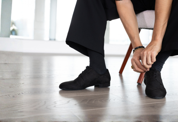 Eine sitzende Person, die die Schnürsenkel ihrer schwarzen Schuhe bindet, während sie eine schwarze Hose und schwarze Socken trägt.