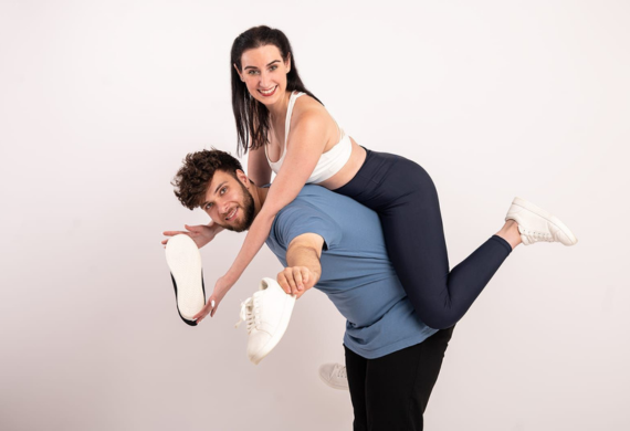 Mann hebt eine lächelnde Frau auf seinen Rücken, beide in legerer Sportkleidung.