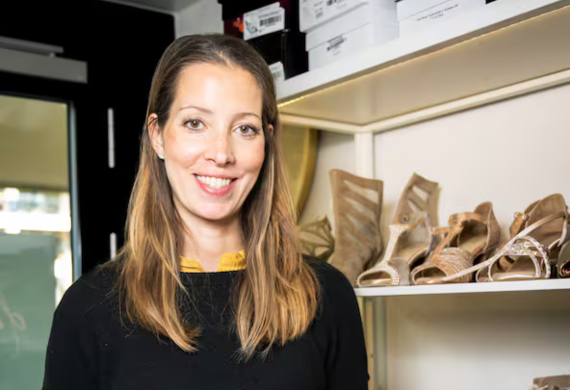 Lächelnde Frau in einem Schuhgeschäft, die neben einem Regal steht, in dem verschiedene hochhackige Sandalen ausgestellt sind.