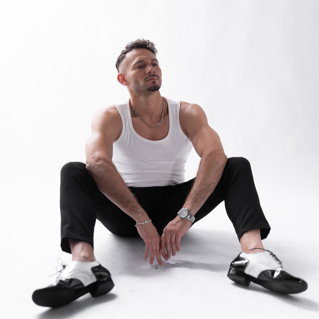 Ein Mann in einem weißen Tanktop und einer schwarzen Hose sitzt vor einem weißen Hintergrund auf dem Boden.